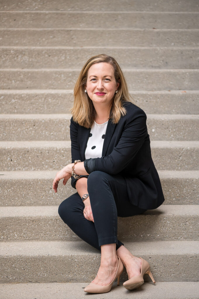 Woman in navy blue suit and nude high heels leans into camera while sitting on white concrete stairs