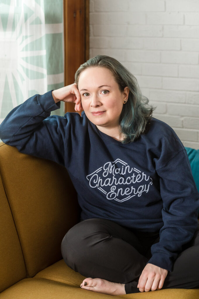 Woman with teal hair wearing sweatshirt that reads "main character energy" as she relaxes on a yellow couch