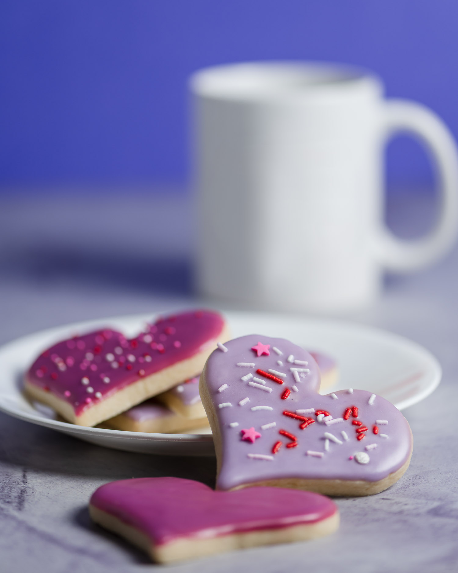 pink and Purple heart shaped sugar cookies are piled on a plate with a mug blurred in the background