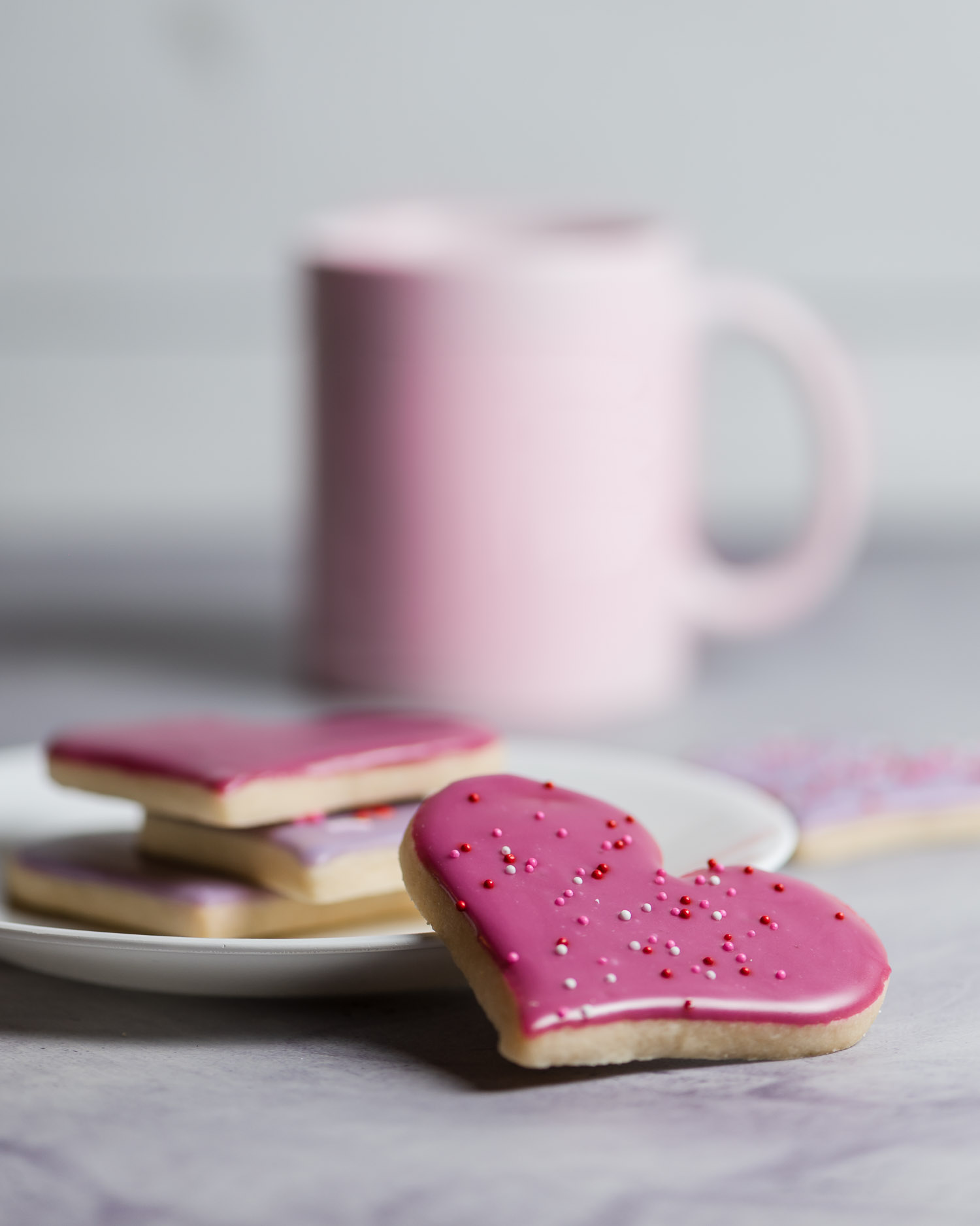 pink and Purple heart shaped sugar cookies are piled on a plate with a mug blurred in the background - after edit