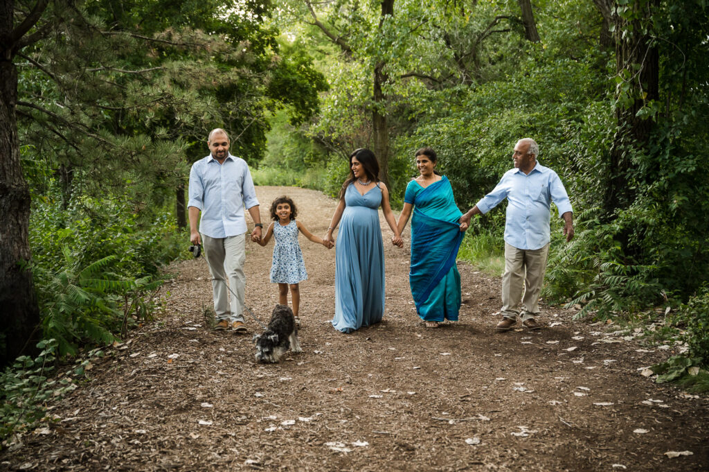 Family along with with grandparents and dog hold hands and walk through forest during a maternity photo session