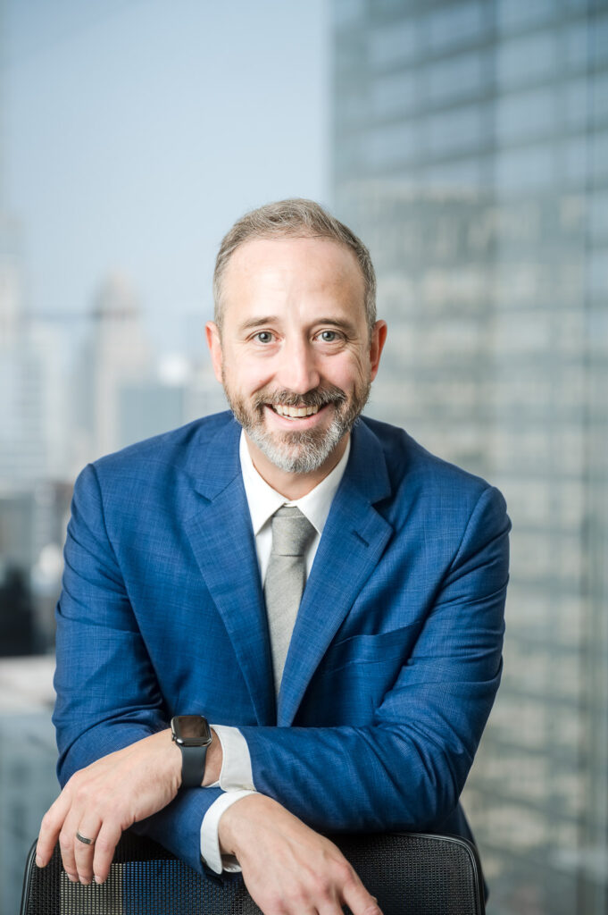 Man in blue suit leans on chair with wrists crossed and smiles at the viewer with the Chicago city visible out the window behind him.