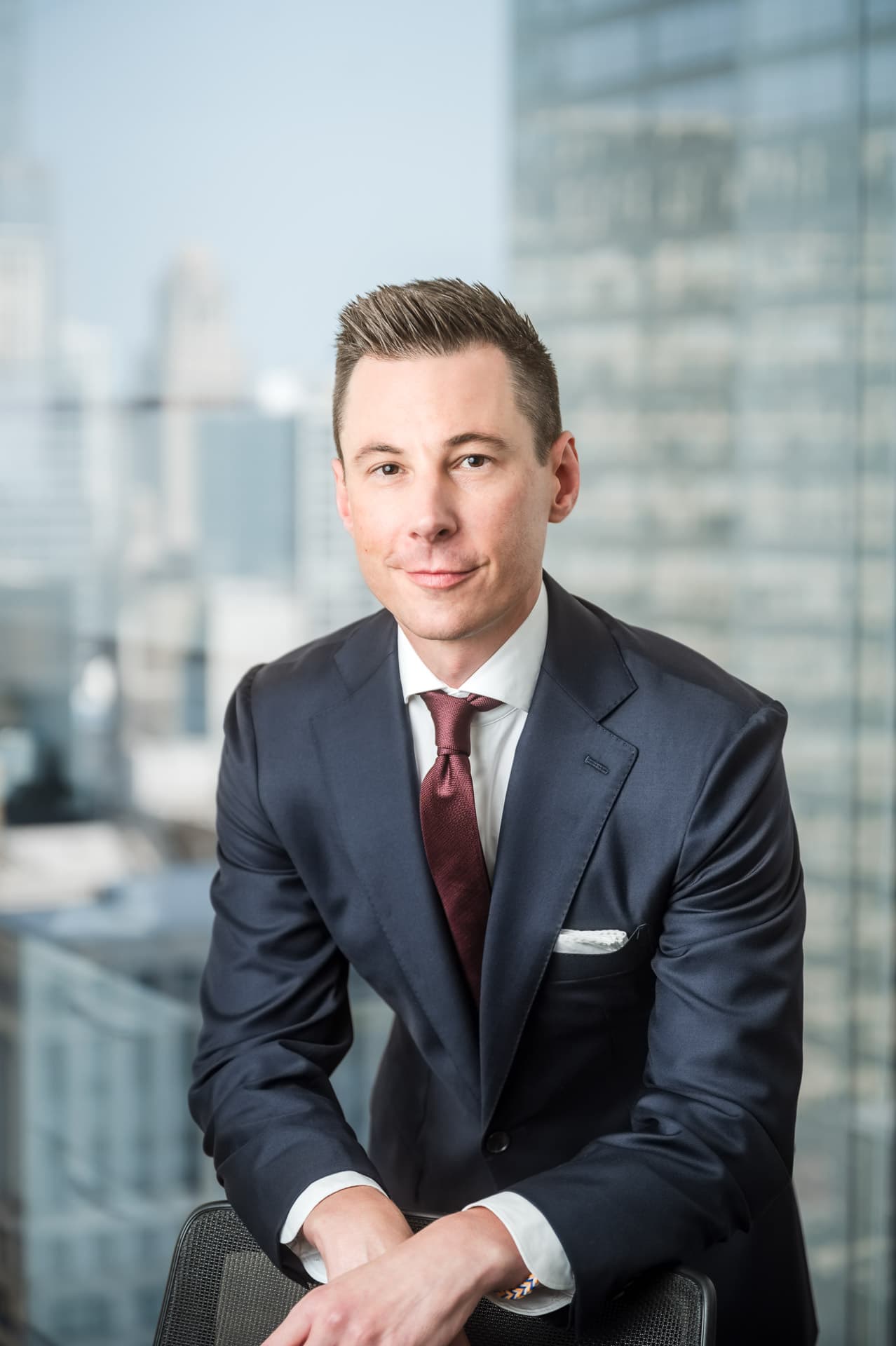 Man in formal suit sits in front of window with Chicago cityscape visible outside