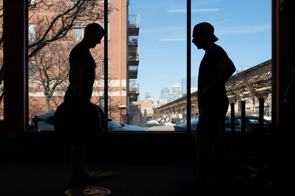 Silhouettes of two men, one is lifting large weights in front of a gym window with a view of the Chicago cityscape out the window.