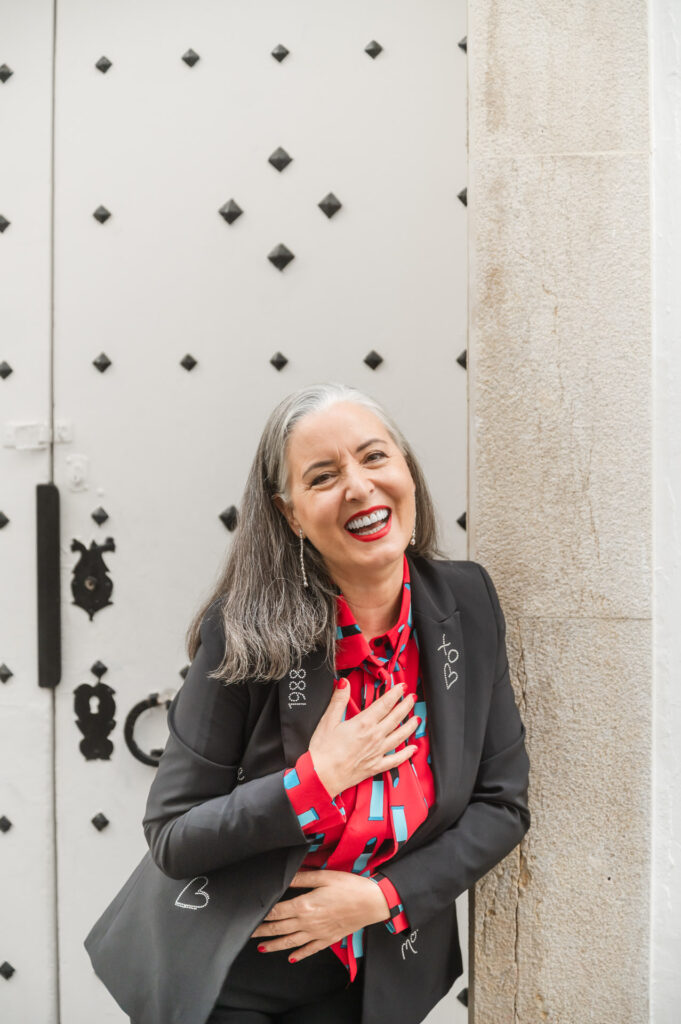 Woman with beautiful long gray hair and wearing a red blouse with lipsticks under a rhinestone embellished black blazer laughs in front of a decorative white doorway during a branding portrait