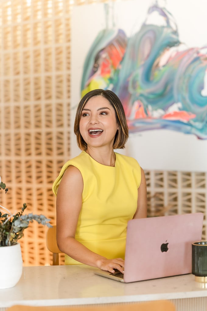 woman in yellow dress laughs while typing at her computer in front of an abstract colorful painting