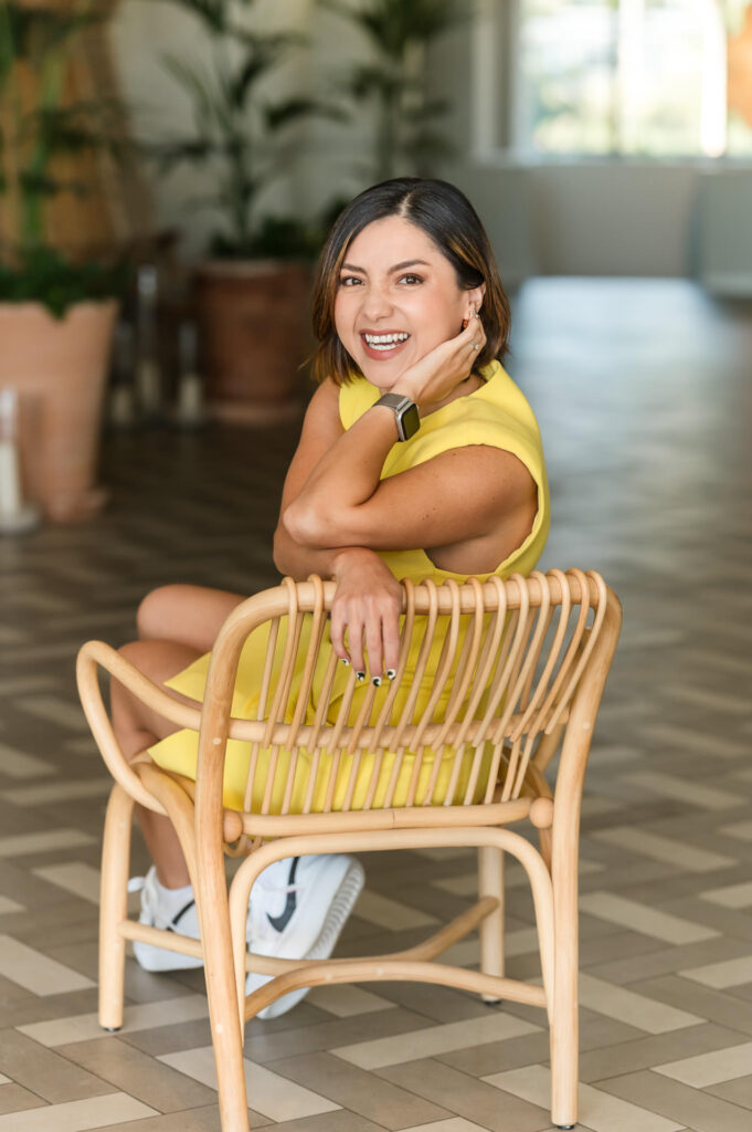 Woman in yellow dress laughs as she tucks her hair behind her ears while sitting in a wicker chair in a large room