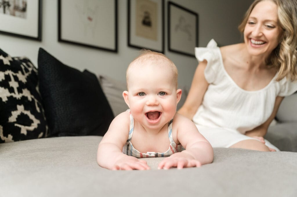 Baby laying on tummy on couch grins at camera with mom smiling behind her.