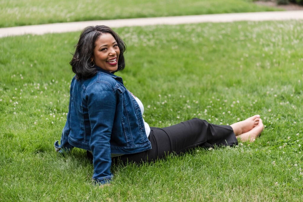 Barefoot woman sitting in grass stretches legs in front of her with ankles crossed and looks over shoulder toward camera and laughs