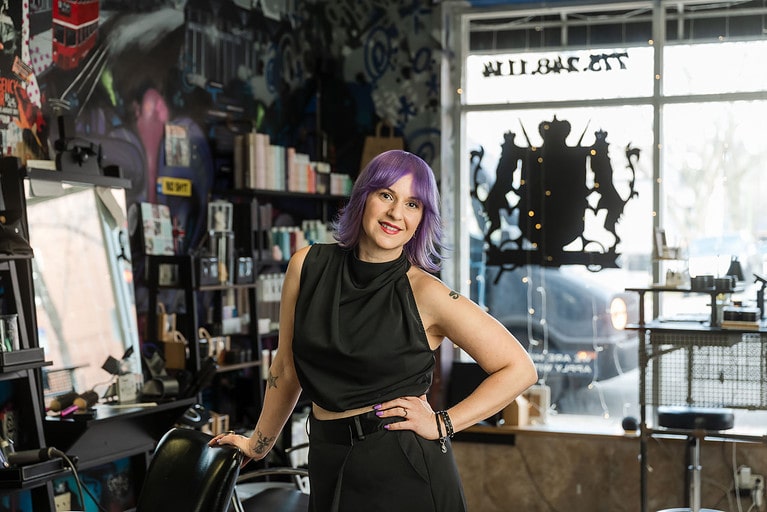 Woman with purple hair leans on hairdressers chair with hand on hip inside her hair salon on Diversey Avenue in Chicago