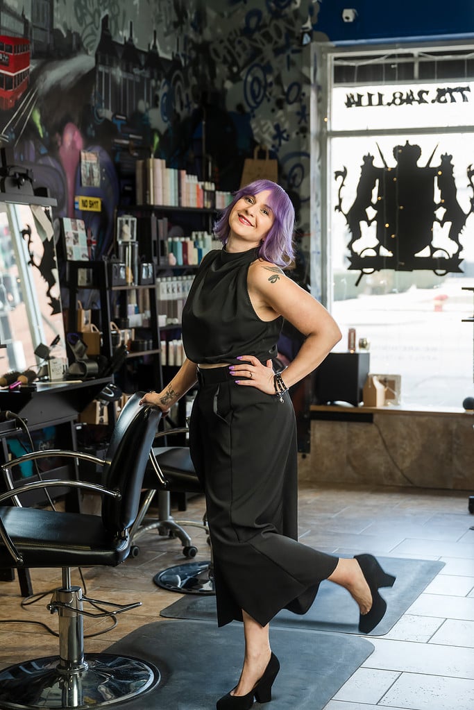 Female stylist with purple hair kicks her foot back in her salon that is a bit grunge and a bit punk rock