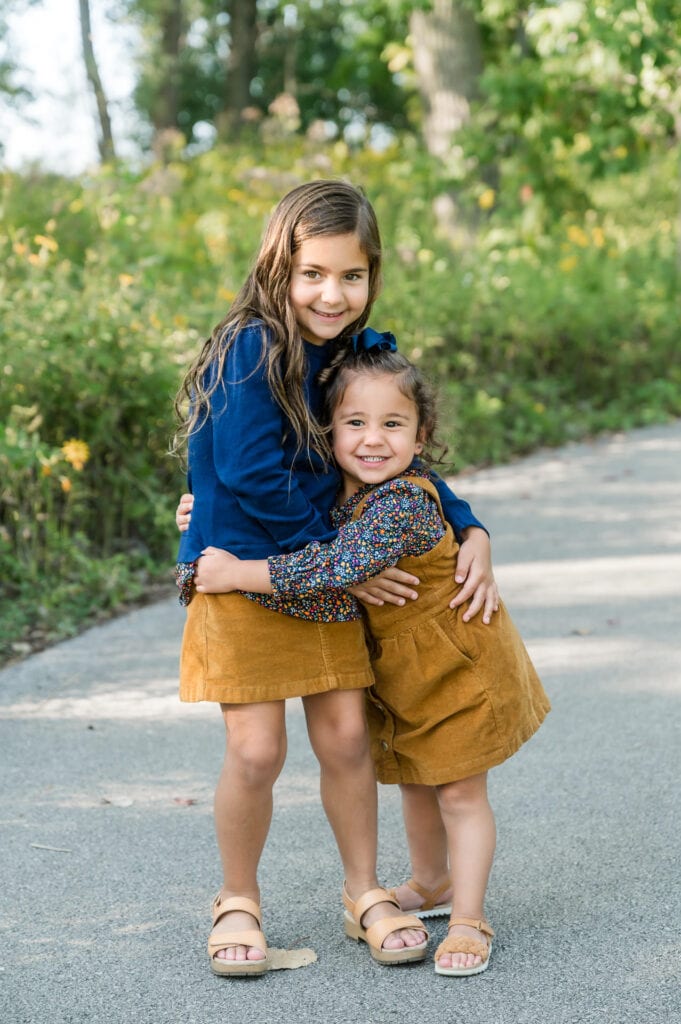 Two young girls wearing navy and mustard give each other a hug in the middle of the path of a nature preserve in Chicago