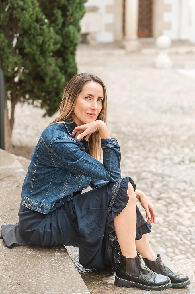Woman in jean jacket, navy blue dress, and black leather boots sits confidently on steps