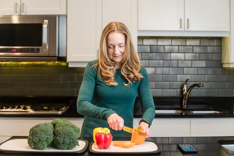 Woman cuts bell peppers, broccoli, and other vegetables in kitchen with black countertop and white cabinets