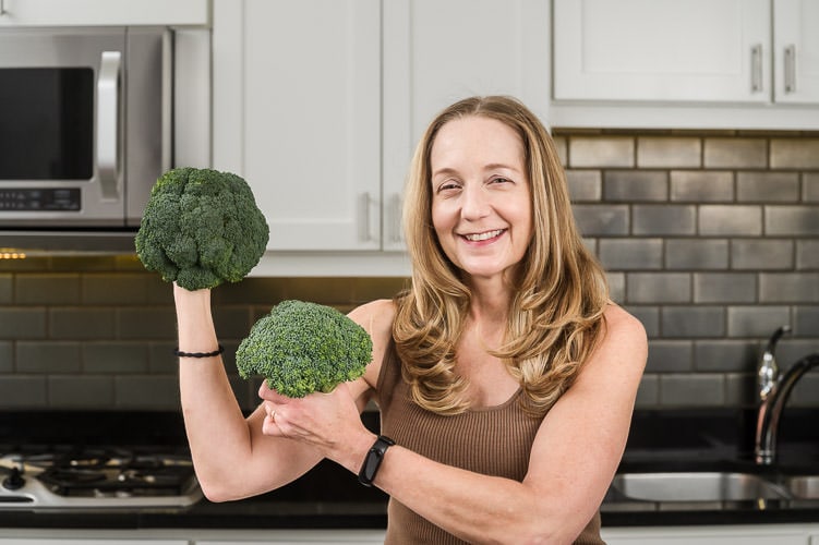 Woman holds broccoli up like gym weights in kitchen