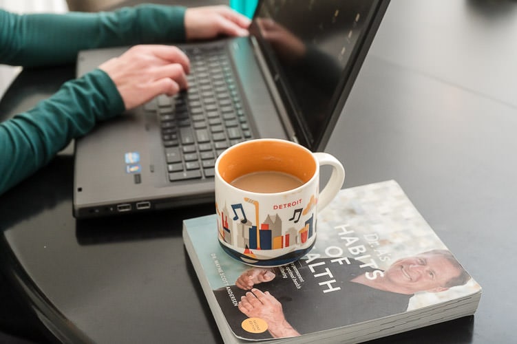 Detail of woman's hands on computer keyboard with a book on health next to her underneath her coffee mug