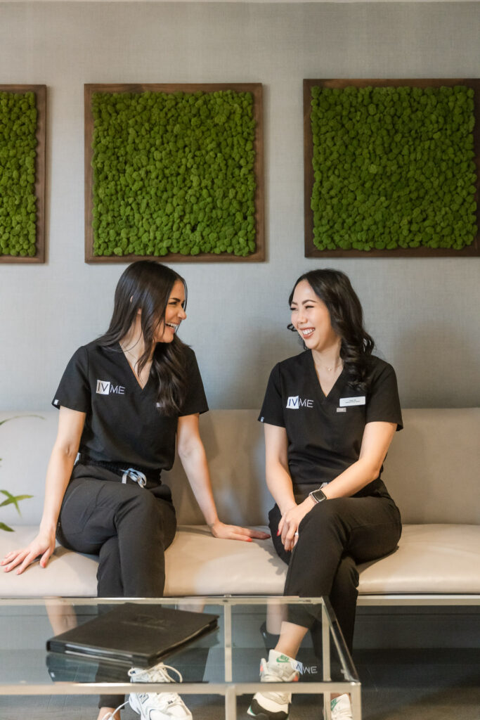 Two women aestheticians sit on a white couch in the waiting room of IVME and laugh together