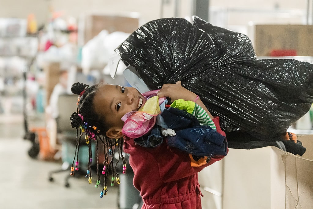 Child working at a volunteer sorting event who just grabbed a huge bag of clothing and can barely see over it.