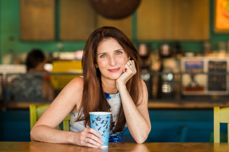 Woman leans on counter of a coffee shop with a cup in hand as she looks thoughtfully into the camera