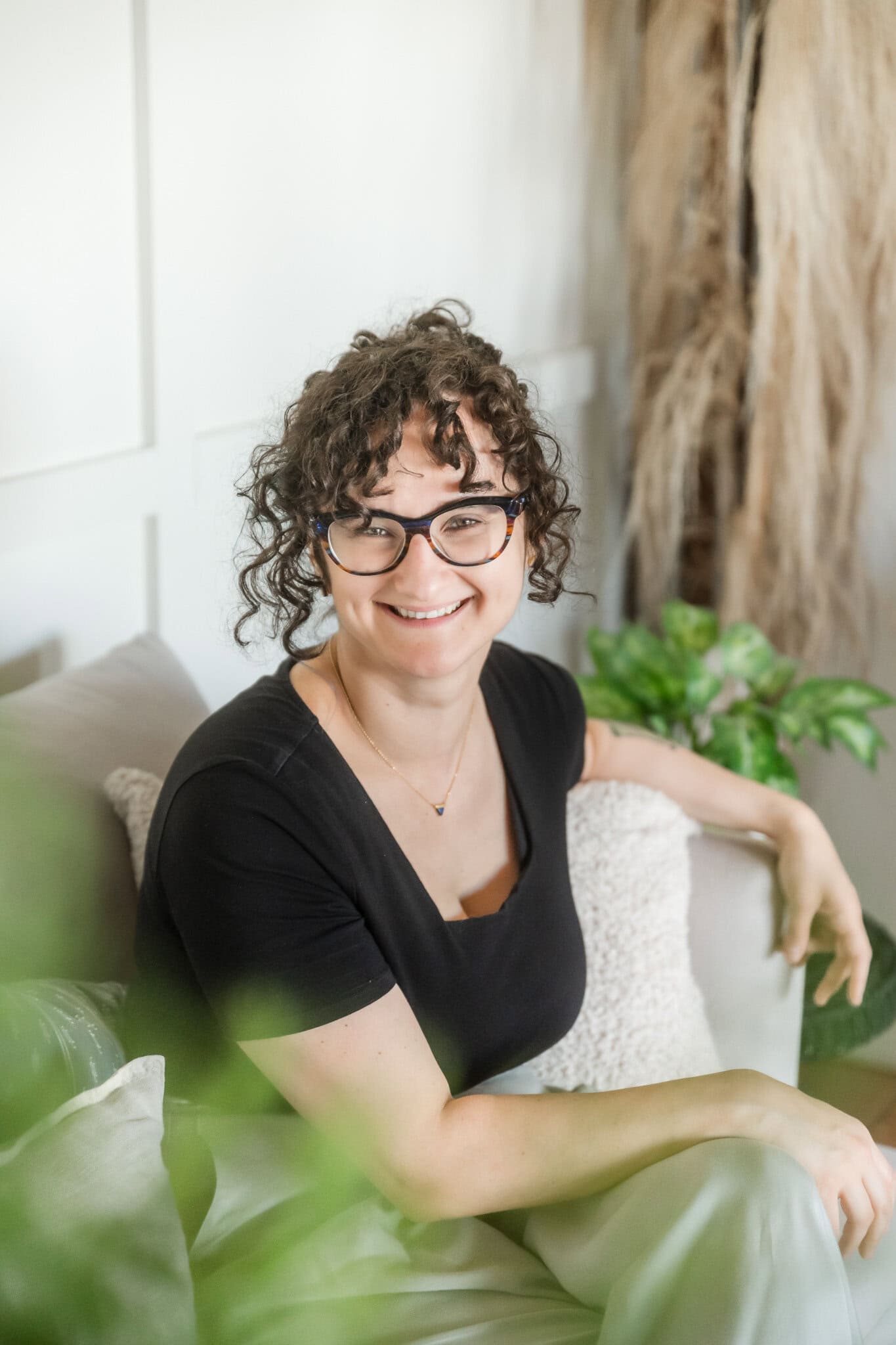 woman with black framed glasses and dark curly hair wearing a black t-shirt sits on a white couch and is surrounded by green plants during a personal branding photography session