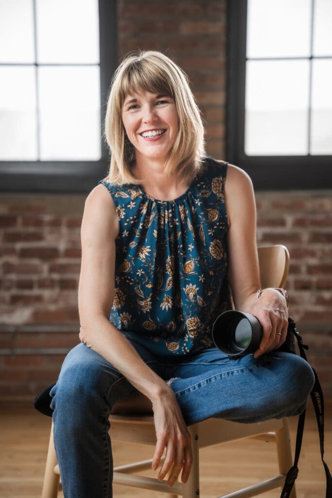 Portrait of Maija Martin Photography sitting on a stool holding Nikon camera in a studio with a a brick backdrop in Chicago