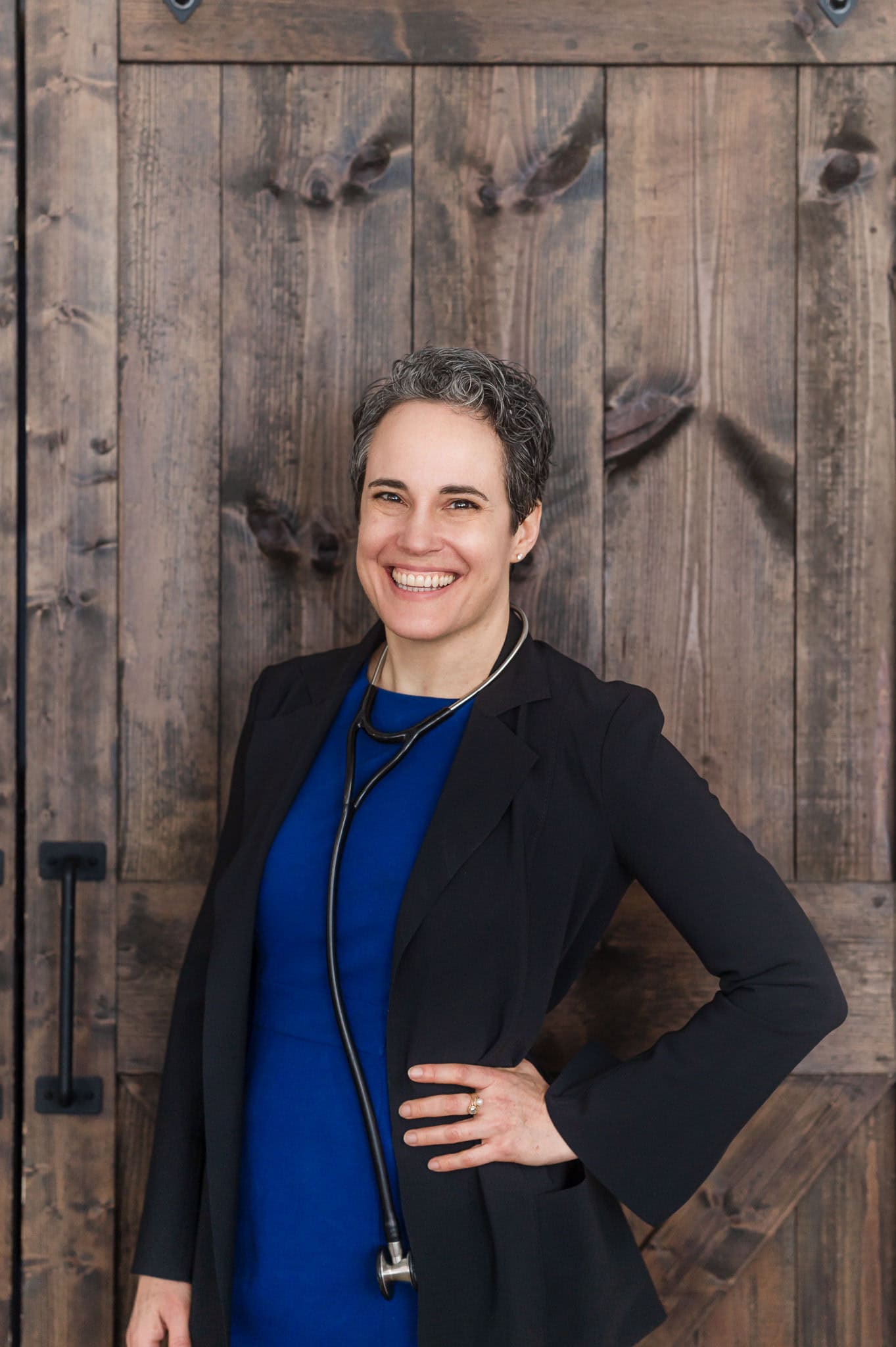 Woman with a pixie style hair cut and wearing a blue dress with a black blazer and a stethoscope leans on wooden door.