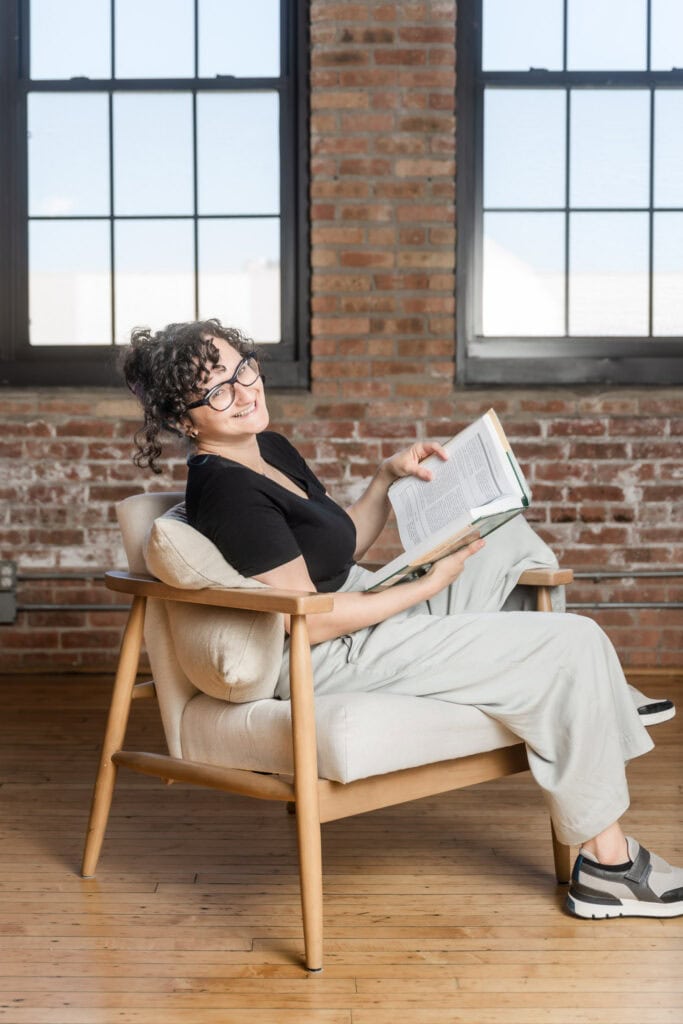 Woman in chair reads book on chinese medicine as she looks back at the camera and laughs