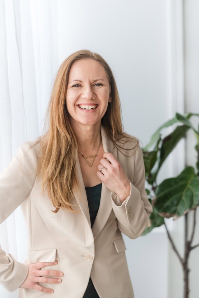 Woman in teal with a tan jacket leans into the camera and laughs