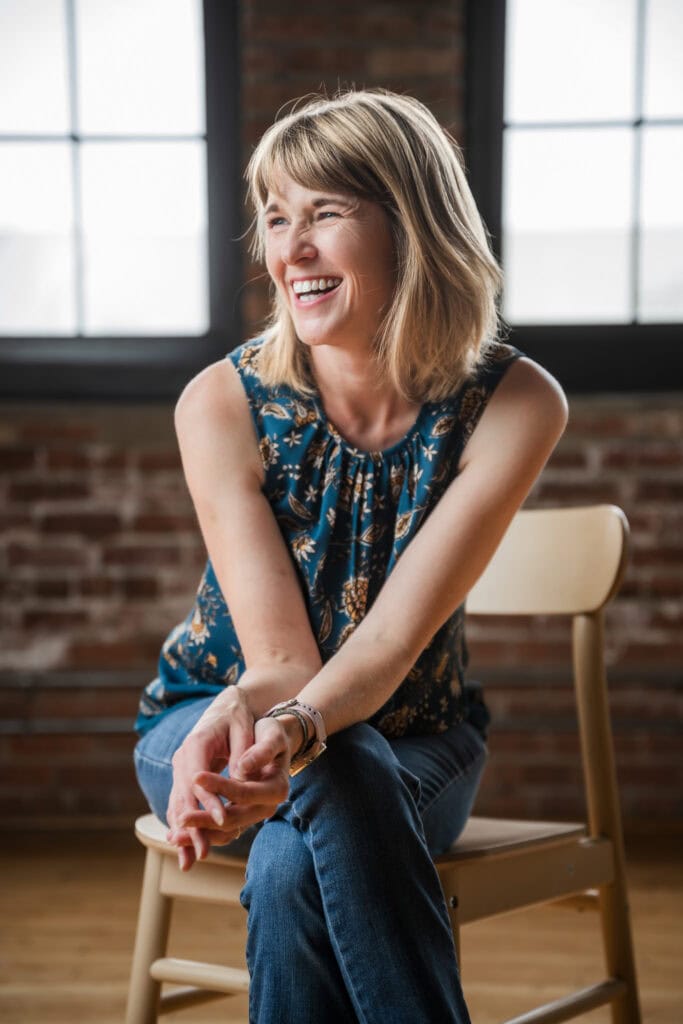 A woman wearing a teal top laughs to a person off-camera