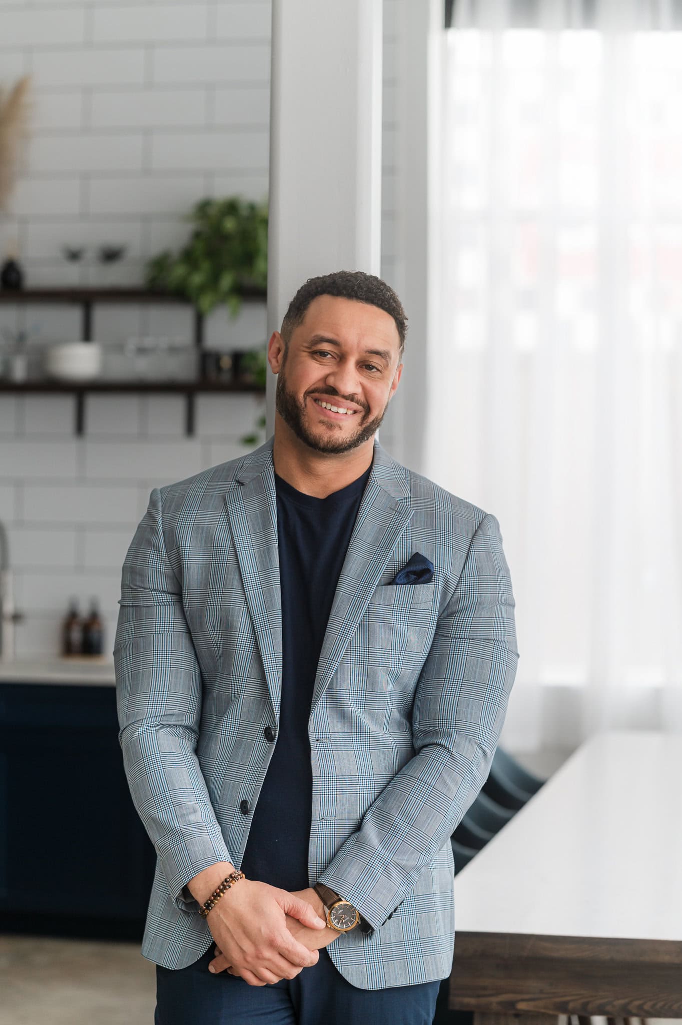 Man wearing a black t-shirt with a blue blazer leans against a white pole with a modern white and blue kitchen space behind him.