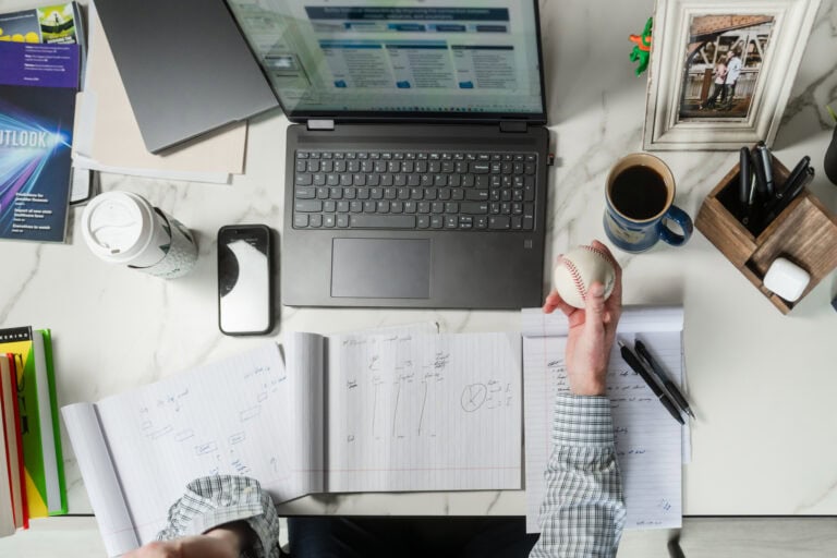 Overview of a man's hand holding a baseball at his desk while working. He's got other artifacts surrounding him giving clues to his personality: his phone, coffee, notes, books, a framed photo