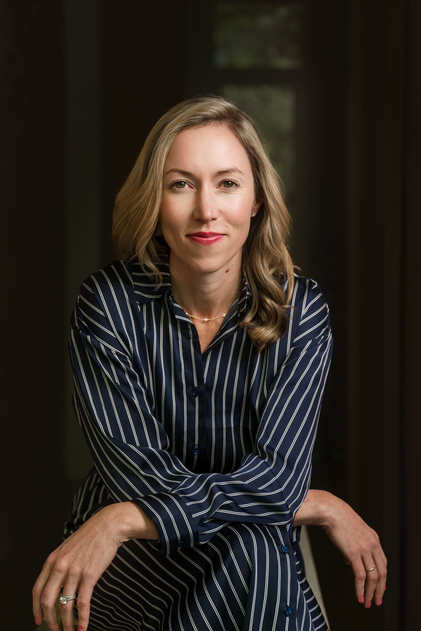 Woman with blonde hair and red lipstick in a navy blue and white striped dress leans into the camera with a more serious, but friendly smile.