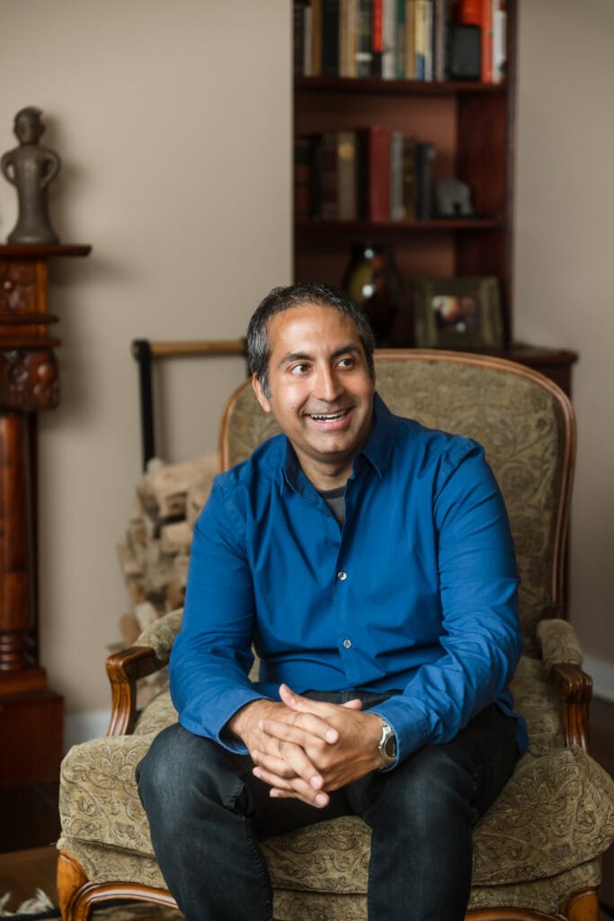 Man wearing a blue button down shirt sitting in a chair looks off camera and laughs