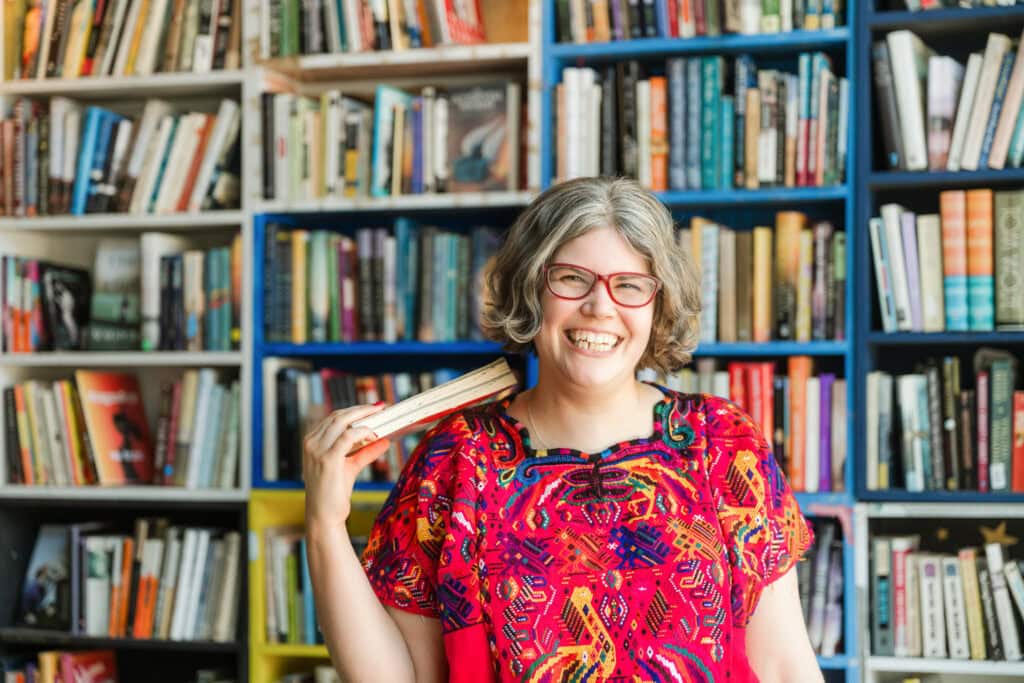 A woman wearing a red top from Guatemala holds a book on her shoulder while standing in front of a bookcase of colorful books