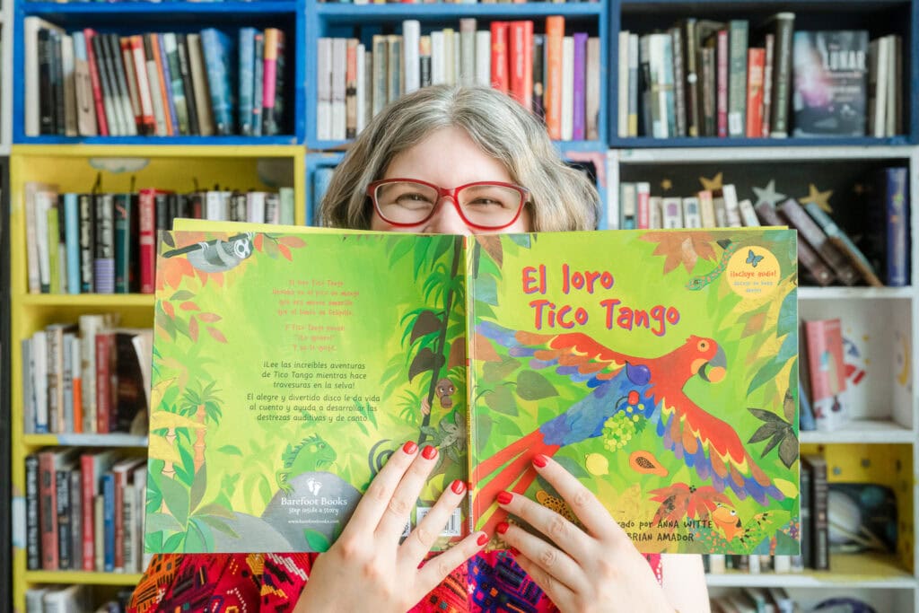 Karla Yatckoske of Uplifting Melodies peeks out from behind a copy of "El loro taco tango" with colorful bookshelves behind her at a bookshop in Pilsen in Chicago.