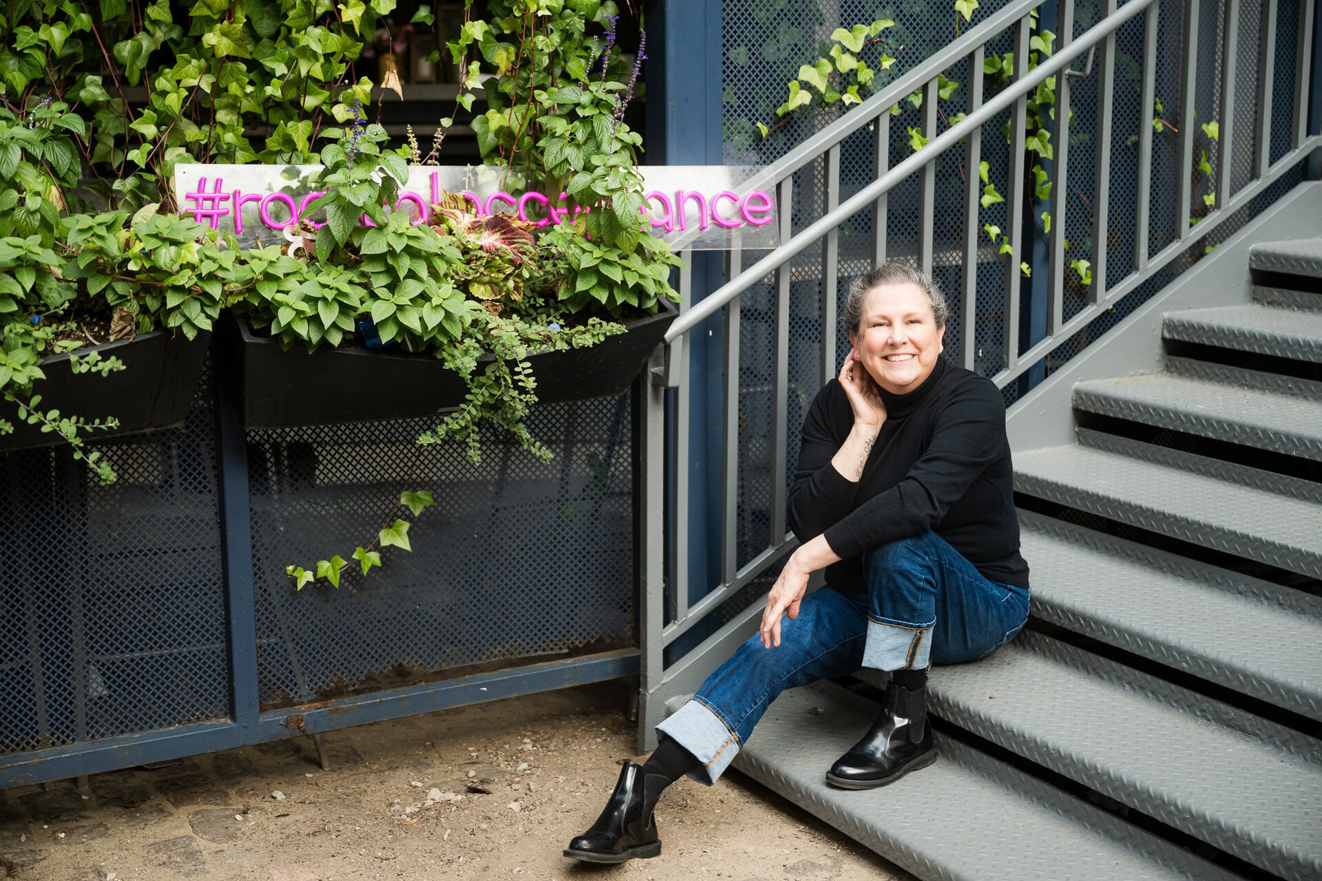 woman sits on a staircase with ivy in the background and a neon sign that reads #radicalacceptance