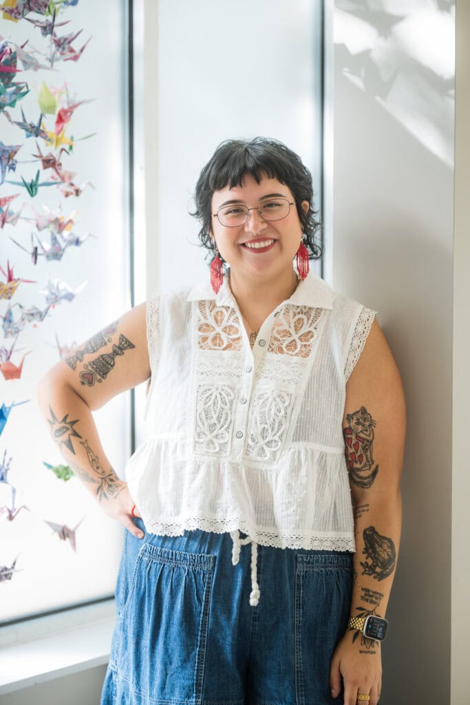woman with dark hair, red earrings, and tattoos leans against a white post with origami cranes to her right.