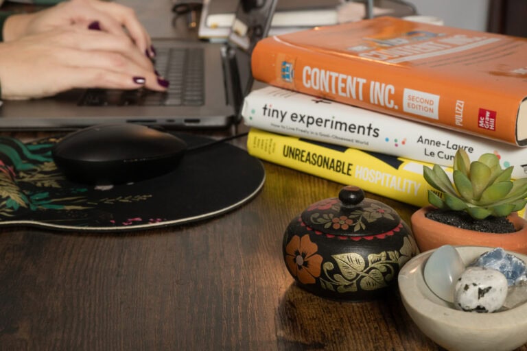 detail of woman's hands with wine colored nail polish typing on a computer behind a stack of books on tiny experiments and content marketing.