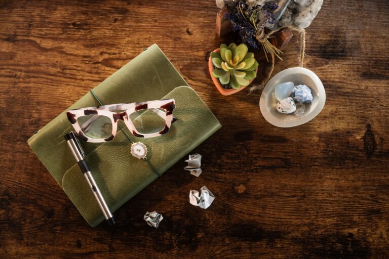 A grouping of objects including glasses, a journal, small plants and crumpled paper against a dark wood backdrop.