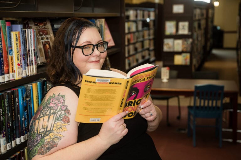 Woman with tattoos on her arm leans on bookcase in library while reading an open book and looking towards the camera