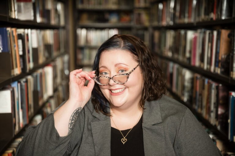 Female librarian peers out over the top of her glasses and smiles at the viewer. She is sitting in front of rows upon rows of books.