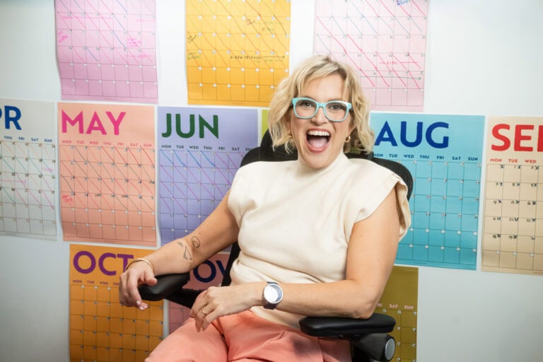 woman with teal glasses sits in desk chair in front of a colorful calendar