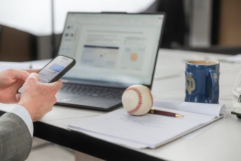 Detail of a man's hands as he texts on his phone with an open laptop in the background showing him mid-work. There's a baseball on the desk next to him with his notepad and pen and a favorite mug from Notre Dame.