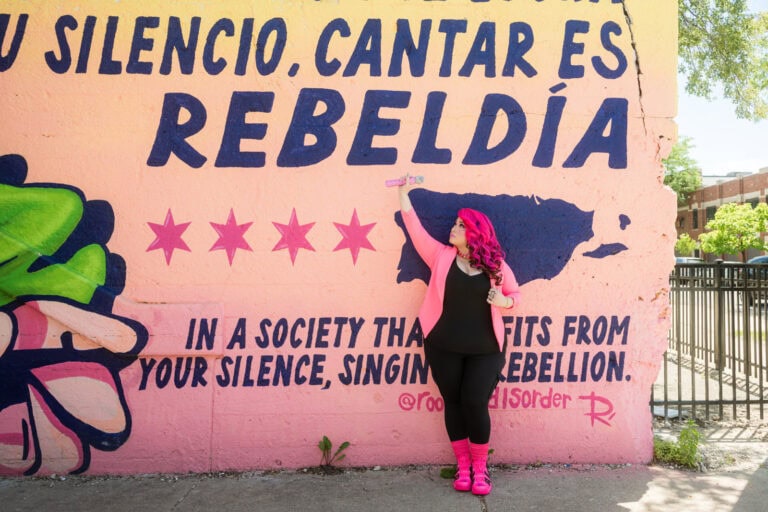 Pinqy Ring, Chicago hip hop artist and educator, leans against a mural with a pink mircrophone raised above her in a rebellion pose. She has bright pink hair, a bright pink jacket, and bright pink shoes on over a black under layer. The mural is mostly pink and is written in Spanish and has the four Chicago stars alongside a navy blue map of Puerto Rico. The message is about not being quiet in a society that tries to make you go silent. To sing is rebellion.